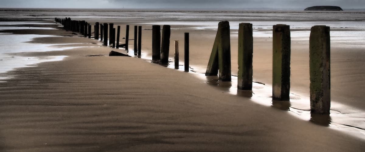 A beach with a wooden groyne and a distant island under a cloudy sky.