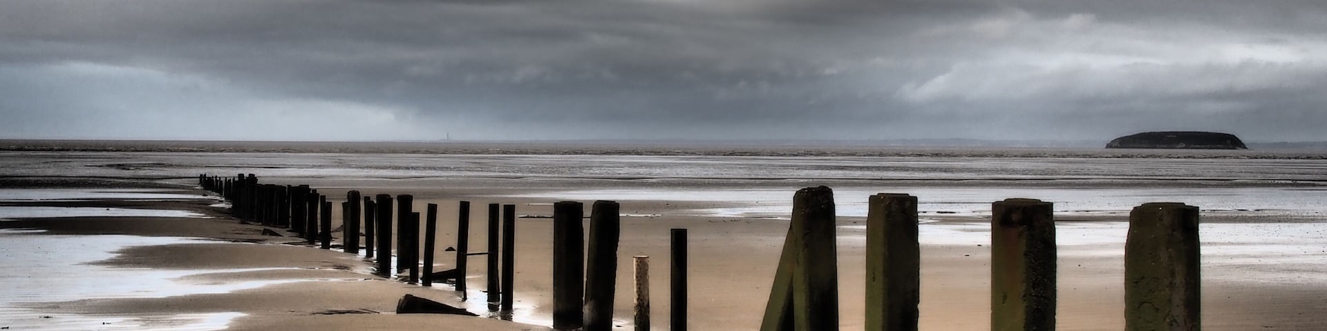 A beach with a wooden groyne and a distant island under a cloudy sky.