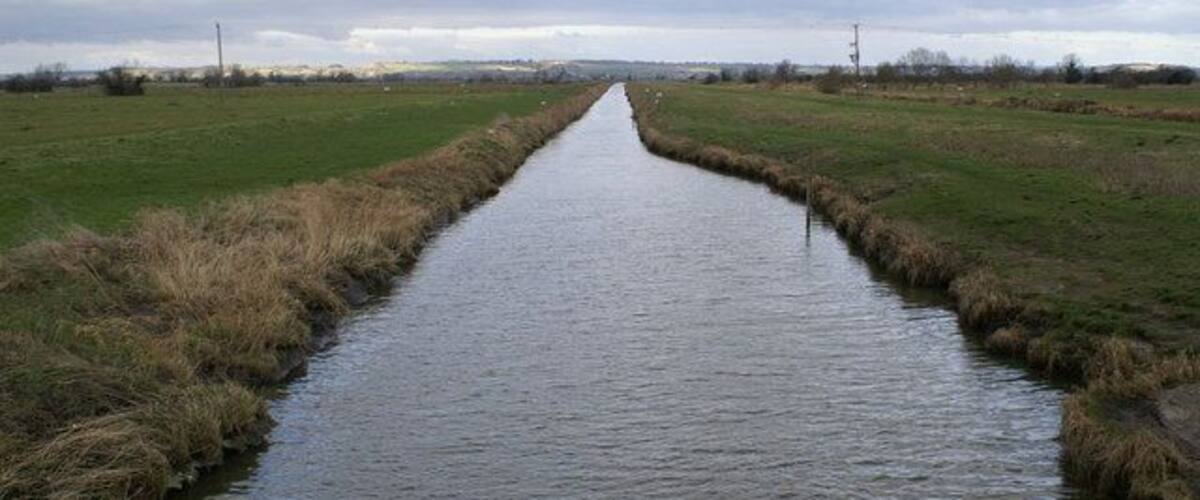 A narrow waterway flanked by grassy fields under a cloudy sky.
