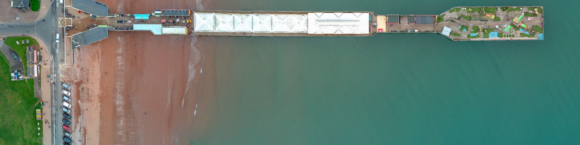 An aerial view of a coastal area with a long pier extending into the water, a parking lot with numerous vehicles, and a small island with buildings and greenery.