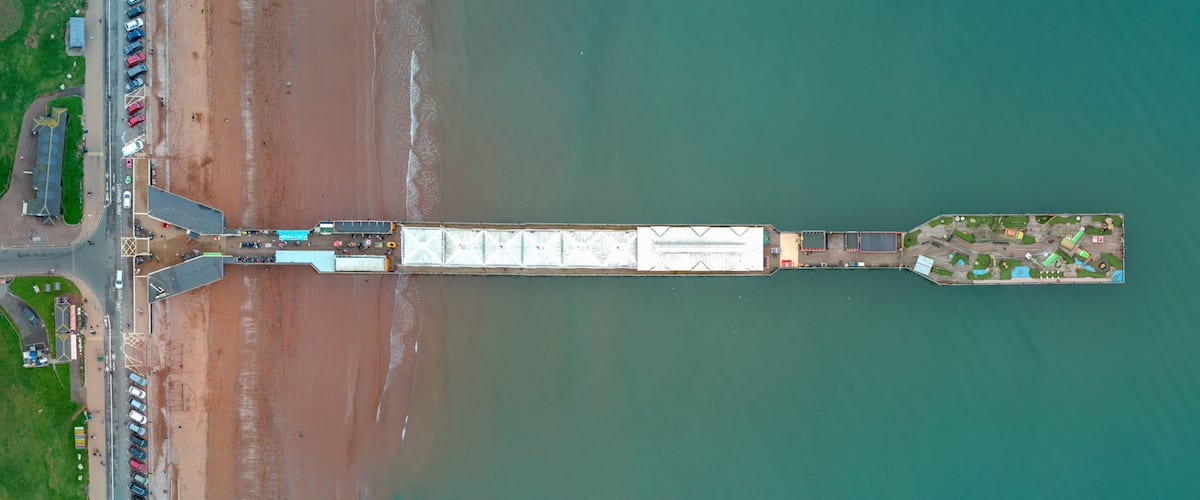 An aerial view of a coastal area with a long pier extending into the water, a parking lot with numerous vehicles, and a small island with buildings and greenery.