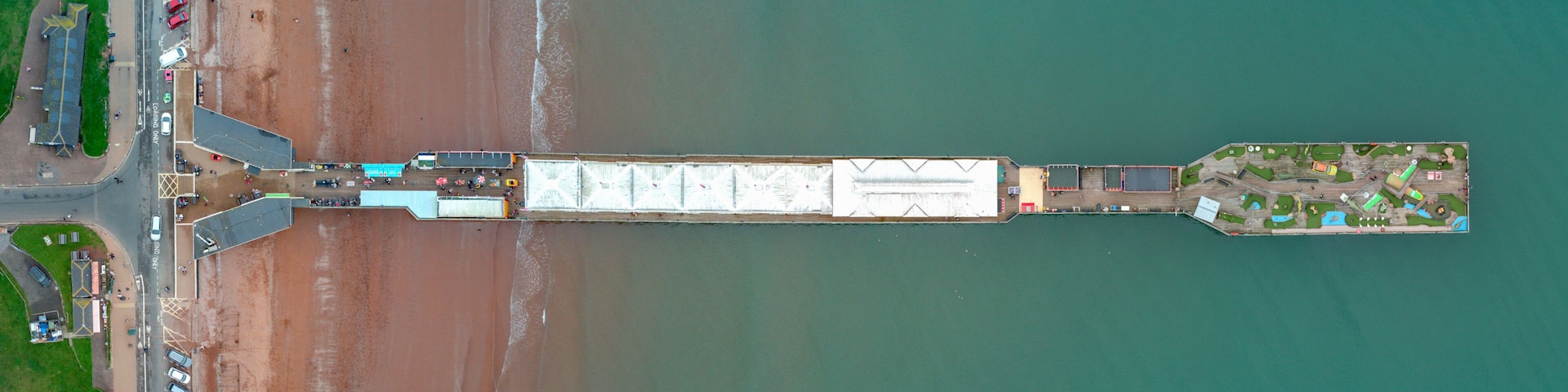 An aerial view of a coastal area with a long pier extending into the water, a parking lot with numerous vehicles, and a small island with buildings and greenery.