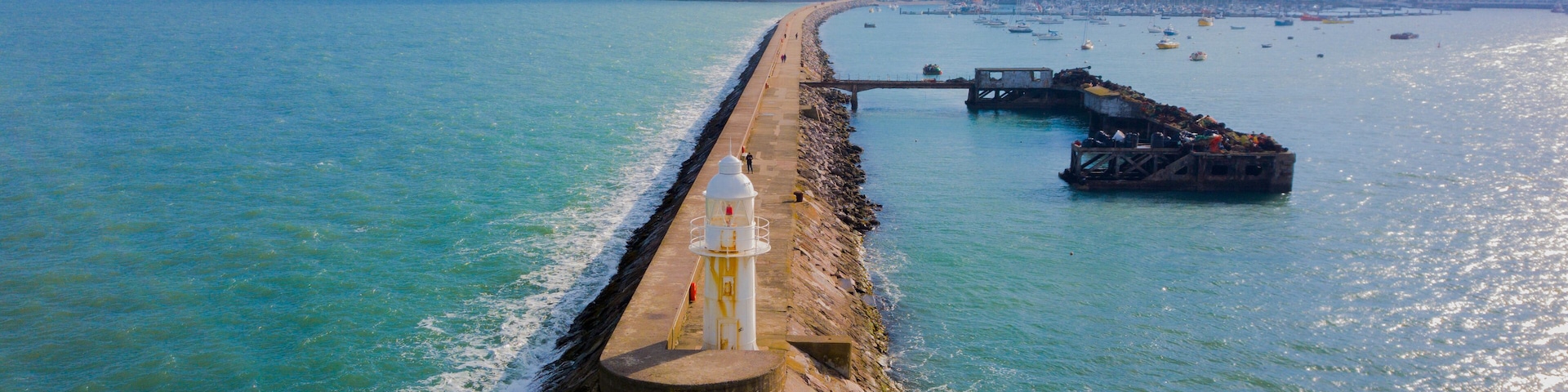 A coastal pier extending into the sea with a lighthouse at the end, a rocky breakwater, and a distant shoreline with buildings.