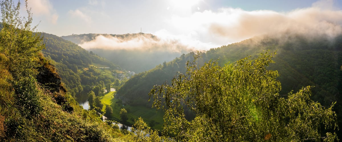 A valley with a river, surrounded by lush green hills and mist-covered mountains.