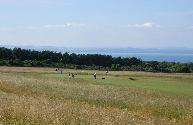 A golf course with players, a grassy foreground, and a forested area with a view of the sea in the background.
