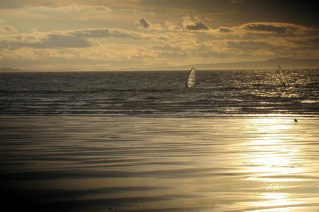 Una playa con un velero en el agua y el sol reflejándose en la arena.