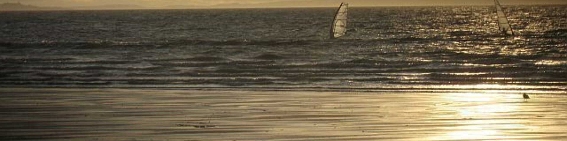 Una playa con un velero en el agua y el sol reflejándose en la arena.