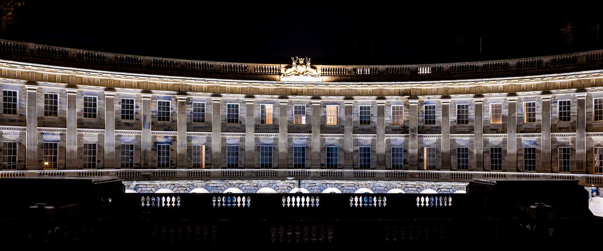 A grand illuminated building with a central statue, large windows, and a reflective pool in front.