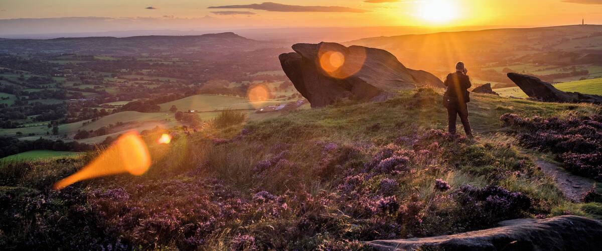 A person taking a photo of a sunset over a hilly landscape with purple heather and large boulders.