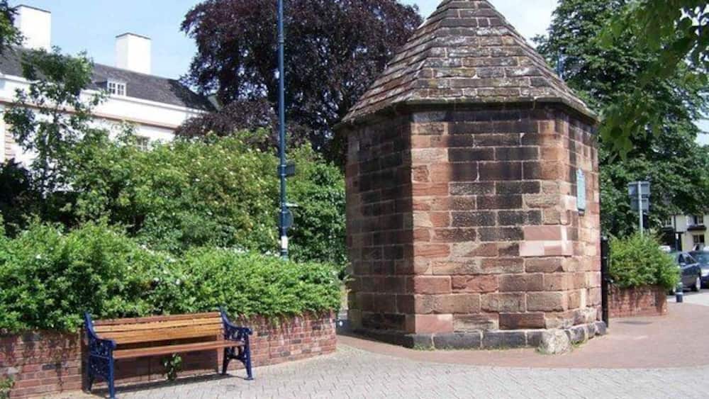 A stone gazebo with a conical roof, a wooden bench, and a brick wall with greenery.