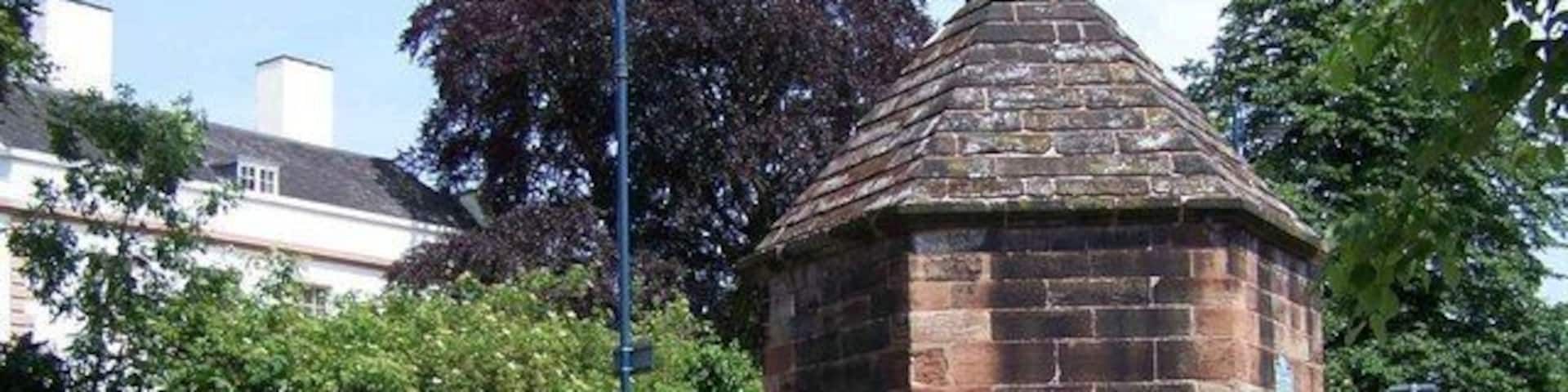 A stone gazebo with a conical roof, a wooden bench, and a brick wall with greenery.
