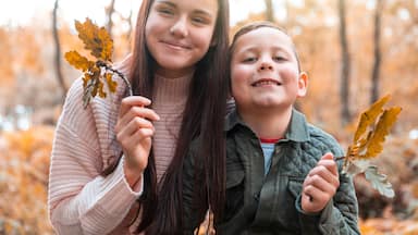 Brother and sister holding dry leaves while standing in Cannock Chase park during autumn