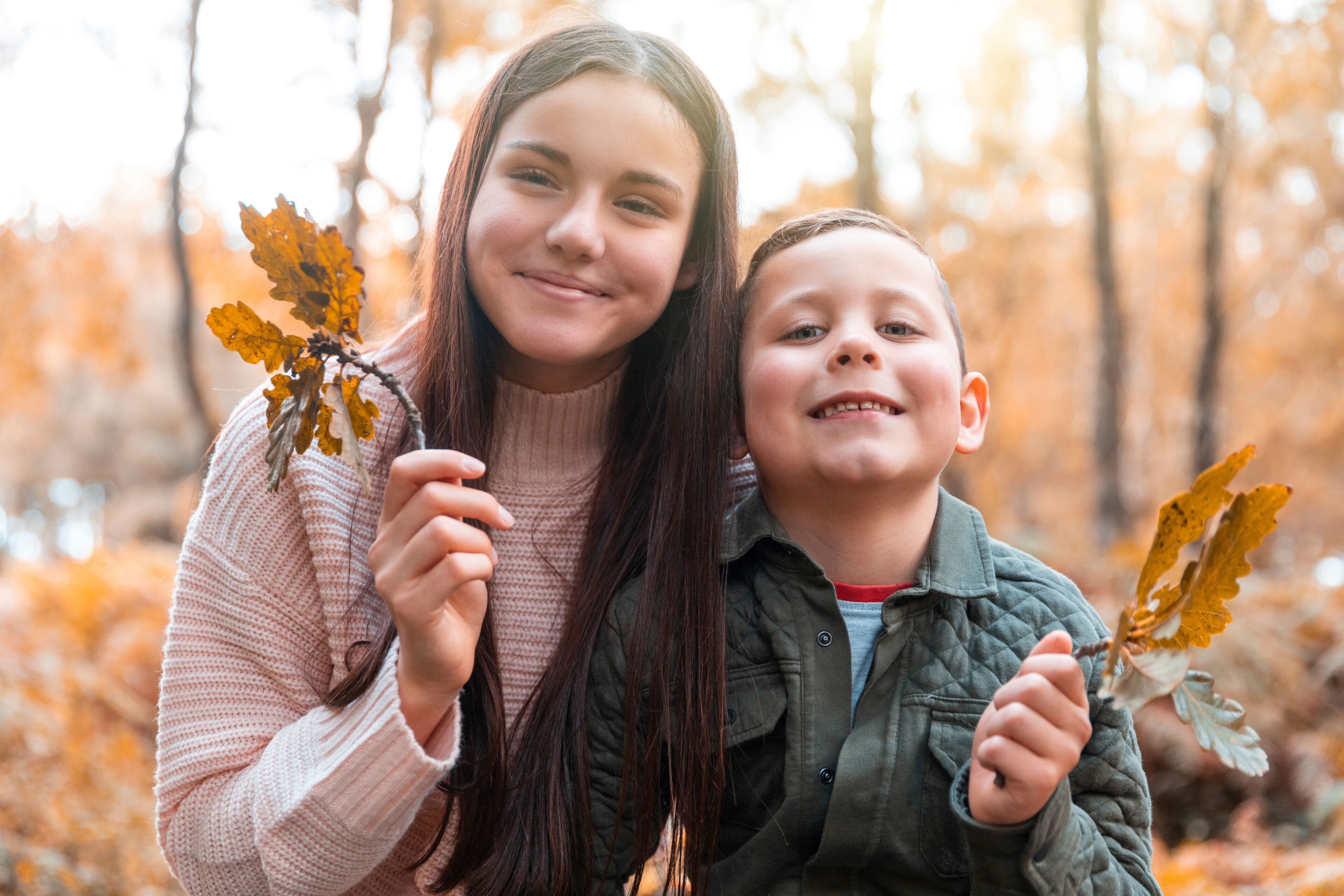 Two individuals holding autumn leaves in a forest with trees in the background.