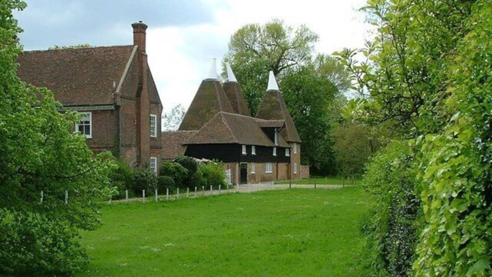 Une maison traditionnelle en briques, avec un toit de chaume et un clocher blanc, entourée d’une végétation luxuriante et d’arbres.