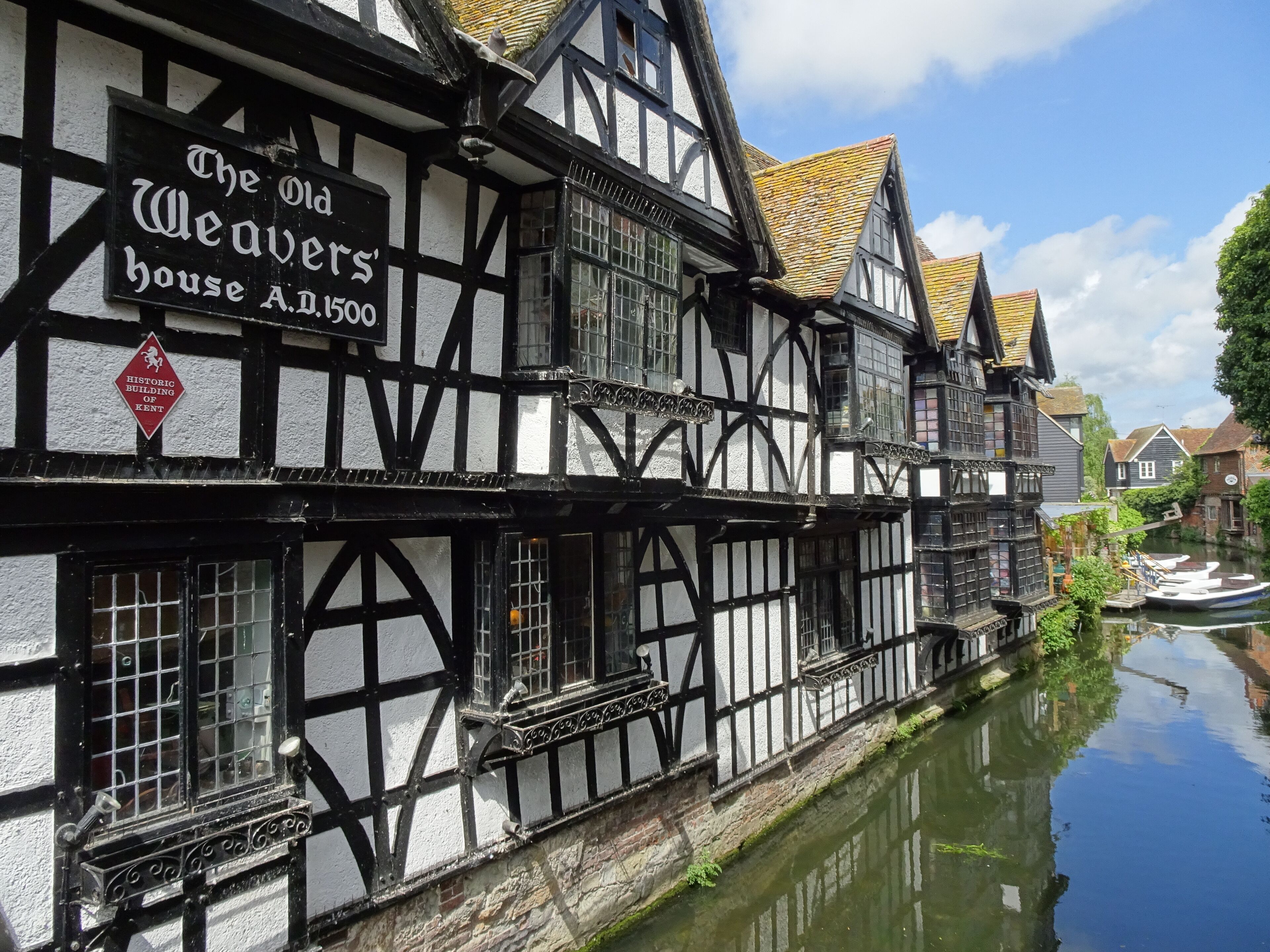 A historic Tudor-style building with black and white timber framing, a sign reading "The Old Weavers' House A.D.1500," and a canal with boats.