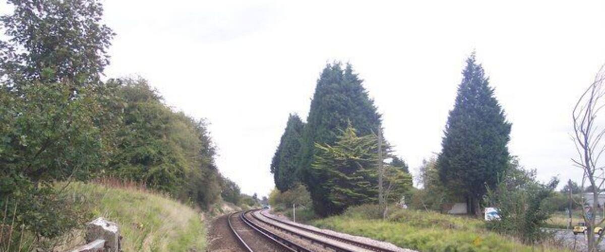 Railway tracks curving through a rural area with trees and a fence on the left.