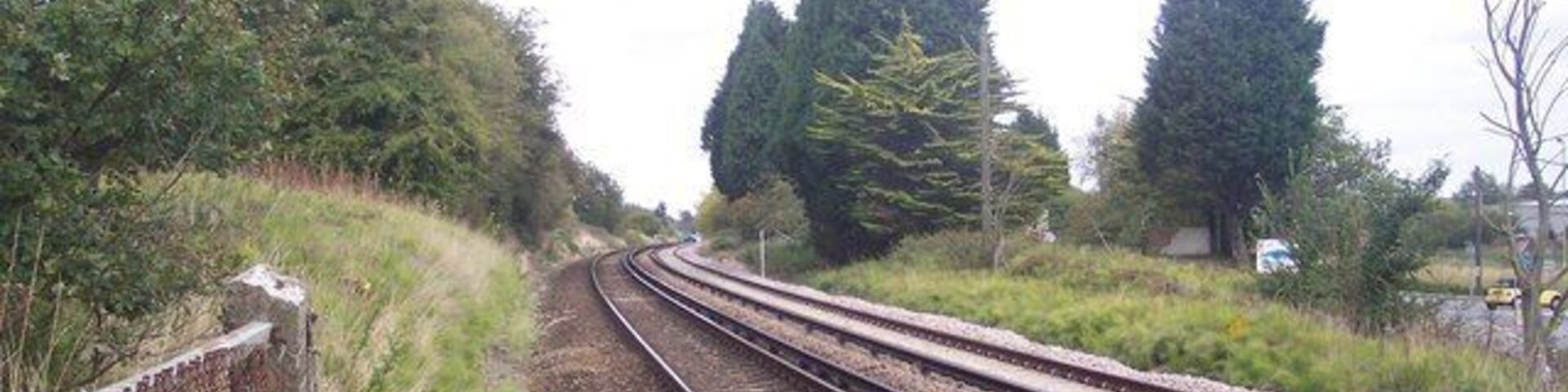 Railway tracks curving through a rural area with trees and a fence on the left.