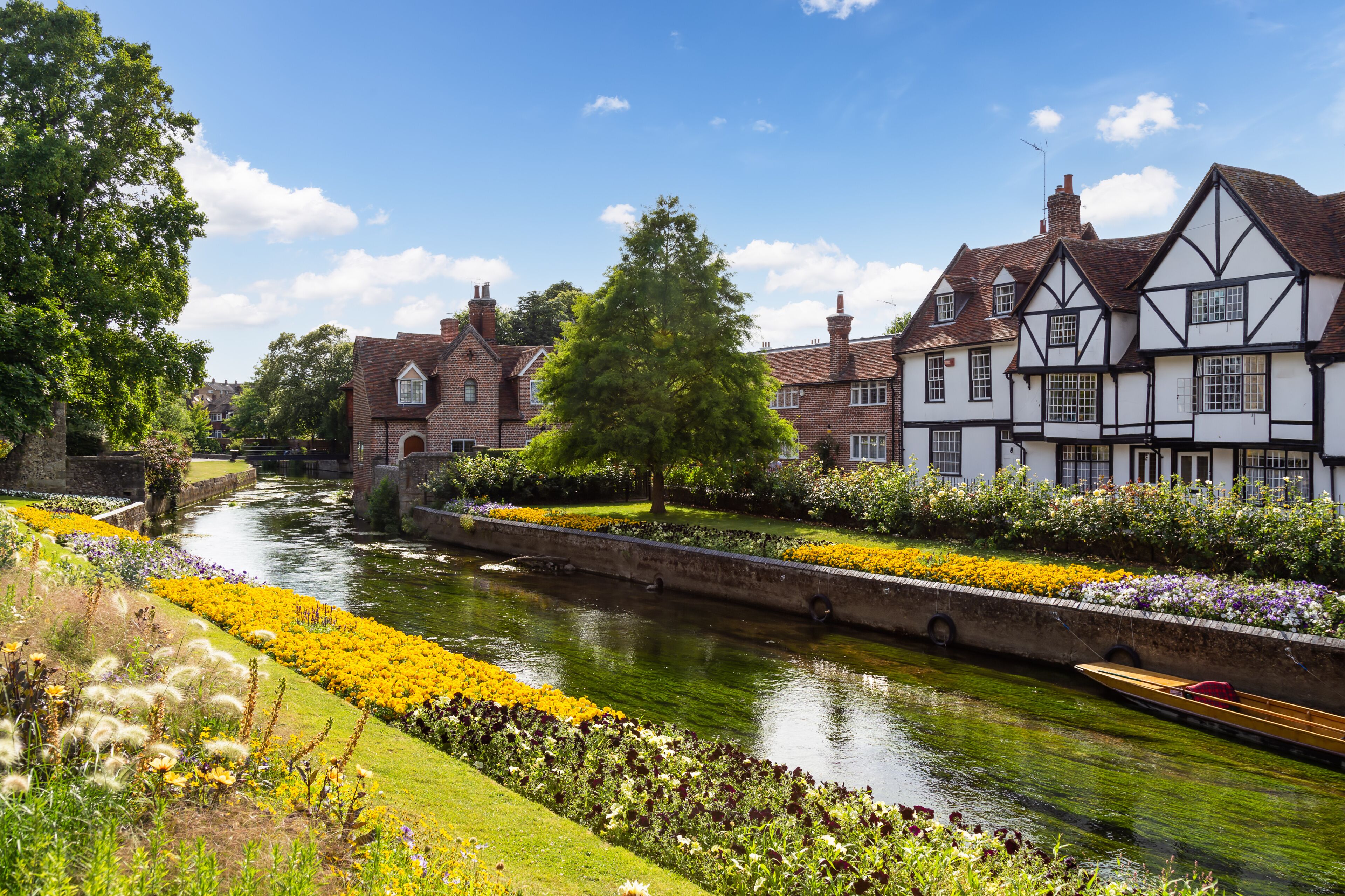 A river with a boat, Tudor-style buildings, and a well-maintained flower garden.