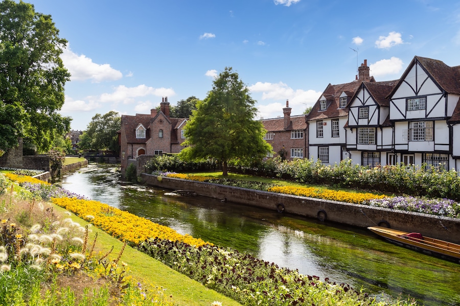 Un río con un bote, edificios de estilo Tudor y un jardín de flores cuidado.
