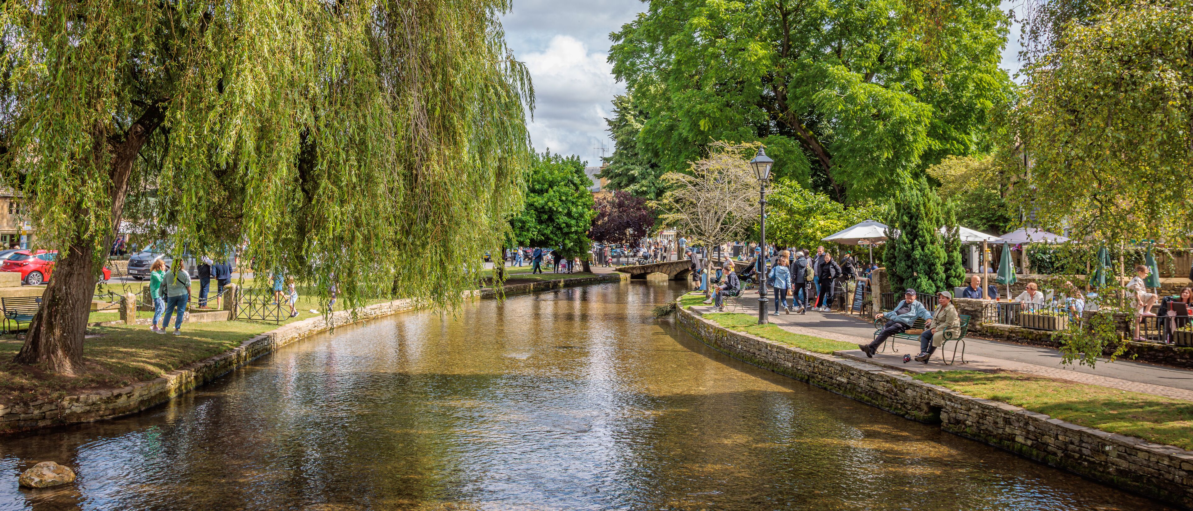 A canal with willow trees, a stone bridge, and people walking and sitting along the path.