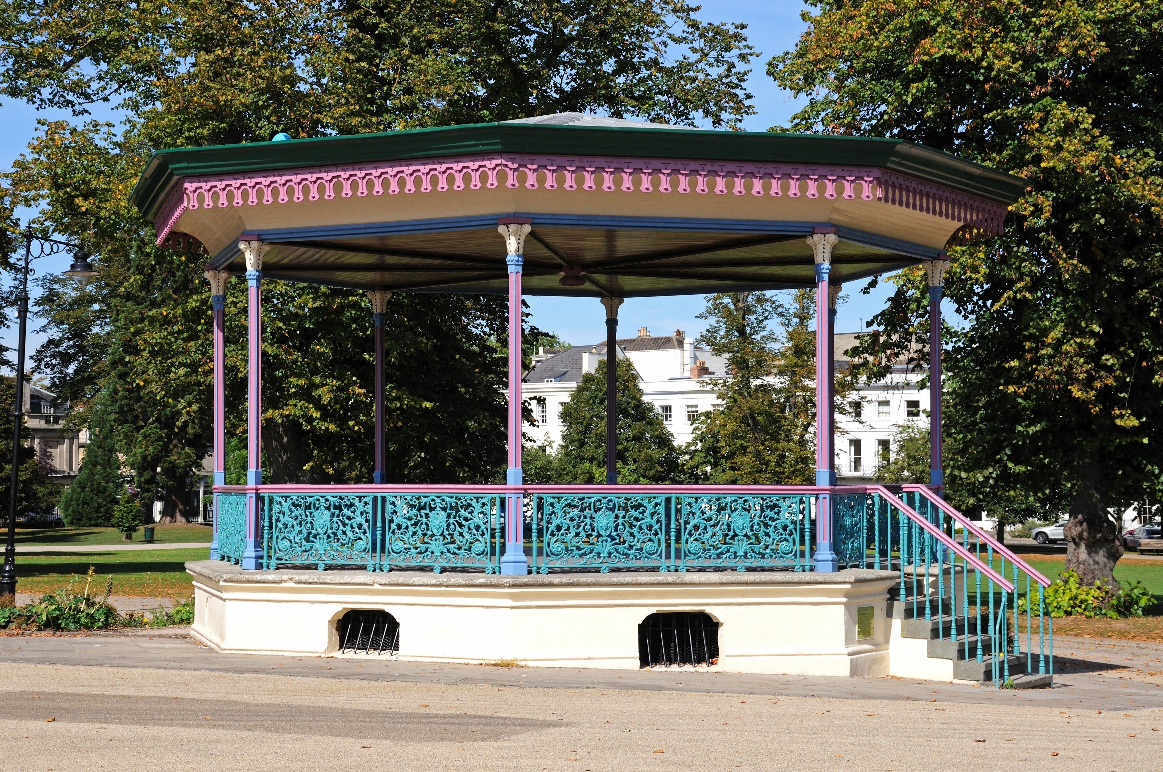 Um gazebo com telhado verde e detalhes rosa, cercado por árvores e um edifício branco ao fundo.
