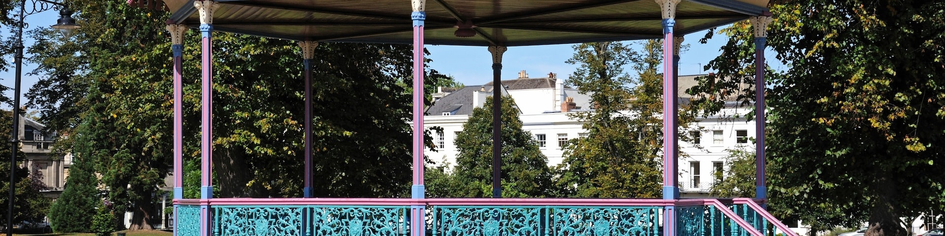 Um gazebo com telhado verde e detalhes rosa, cercado por árvores e um edifício branco ao fundo.