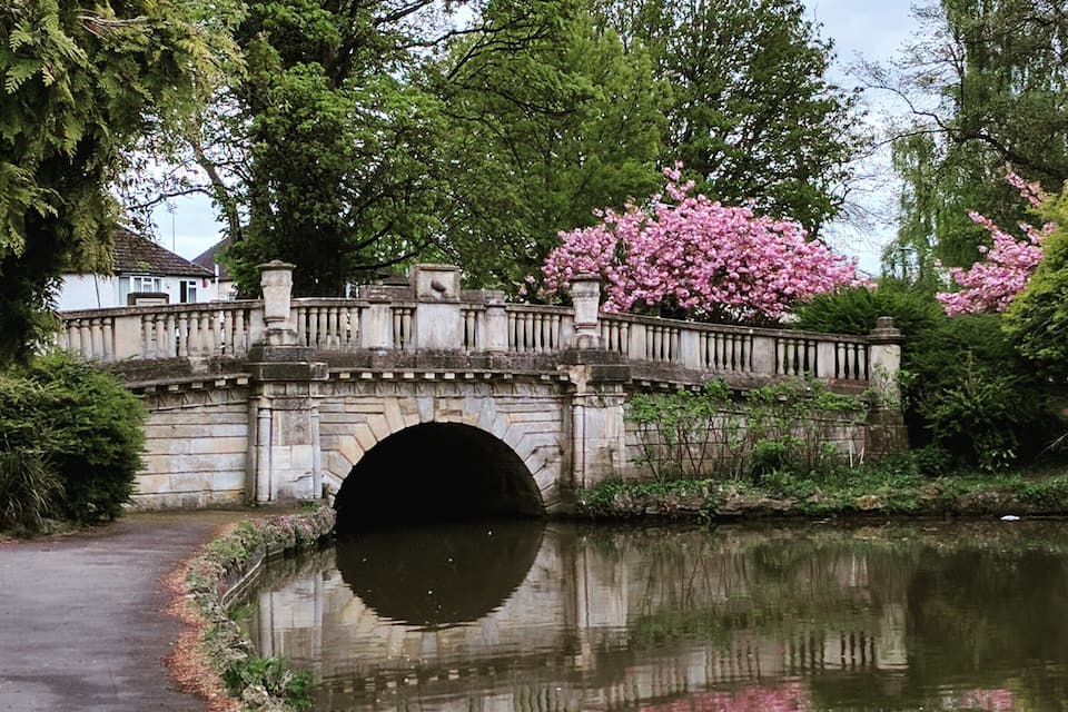 A stone bridge over a canal with a reflection in the water, surrounded by trees and blooming pink flowers.