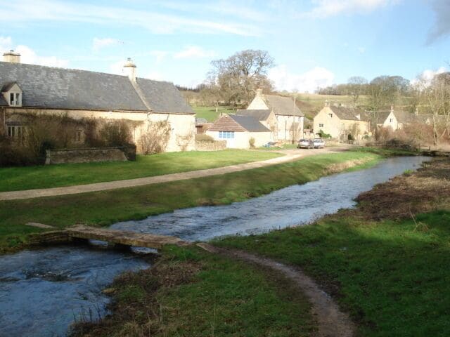 A rural landscape with a narrow stream, stone houses, and a grassy path.