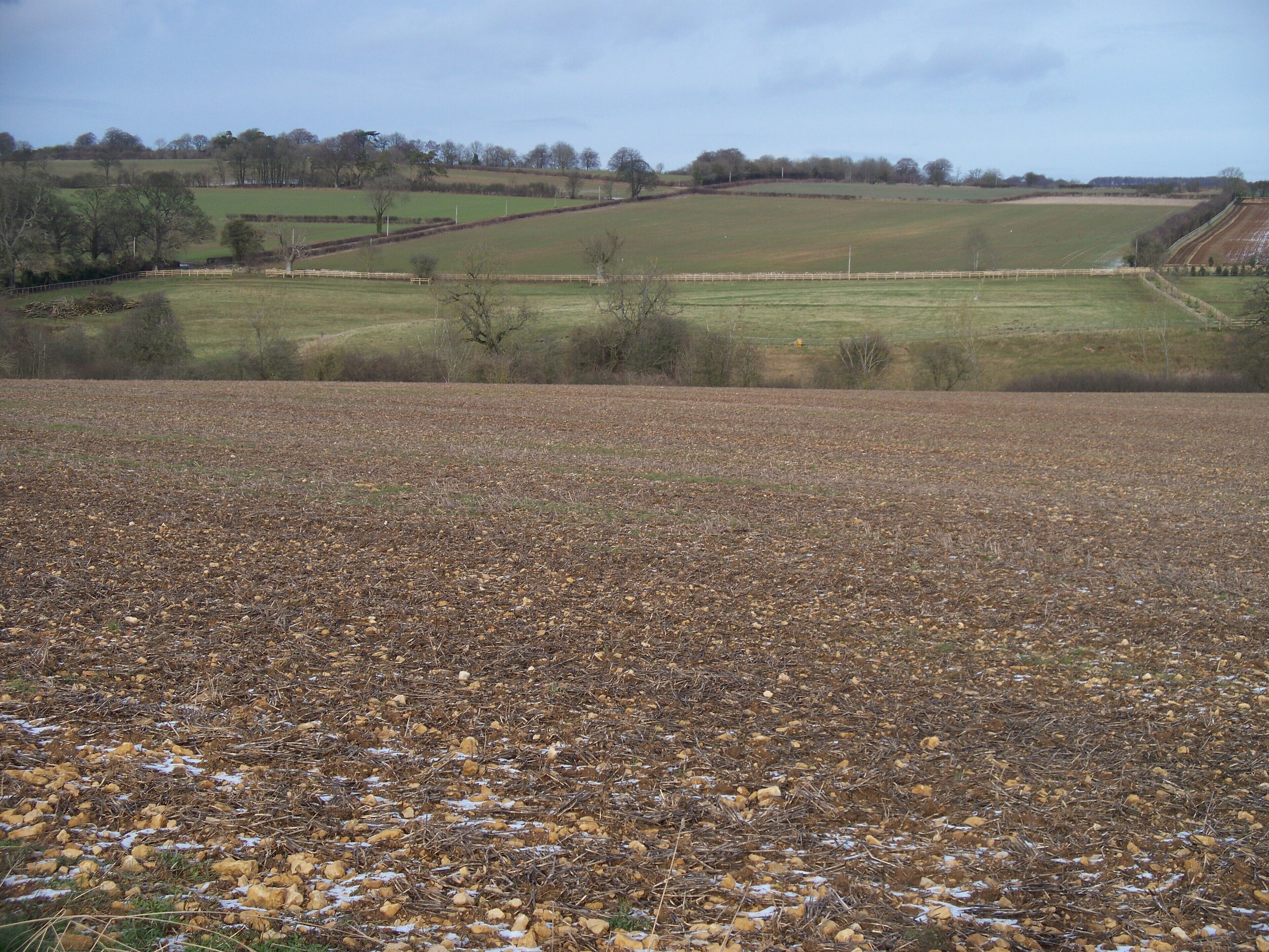 Farmland and lost village This view is looking over the ploughed field and across the valley from the track. The ploughed field appears to be growing a good crop of stones. The first field beyond the trees which mark the course of the stream is the site of a lost medieval village.