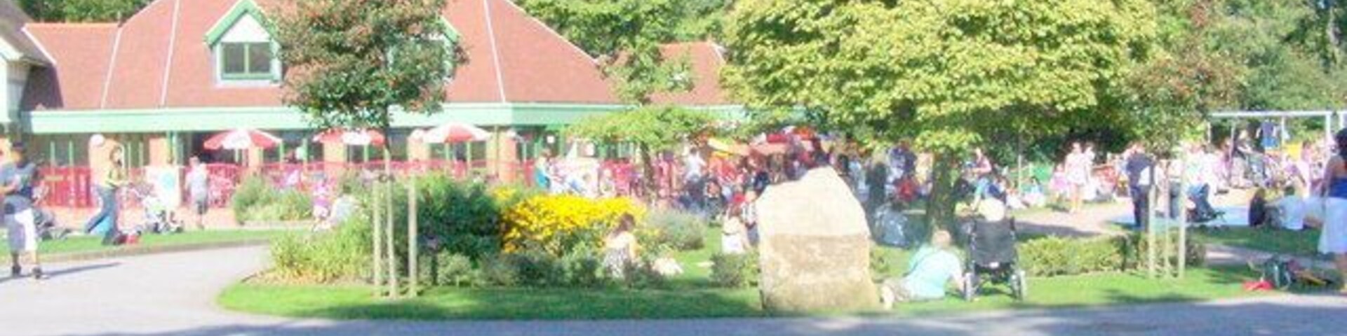 Um parque com uma árvore grande, um edifício com telhado vermelho e pessoas reunidas ao fundo.