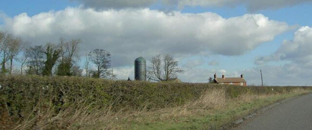 A rural landscape with a road, a silo, and a house in the distance under a partly cloudy sky.