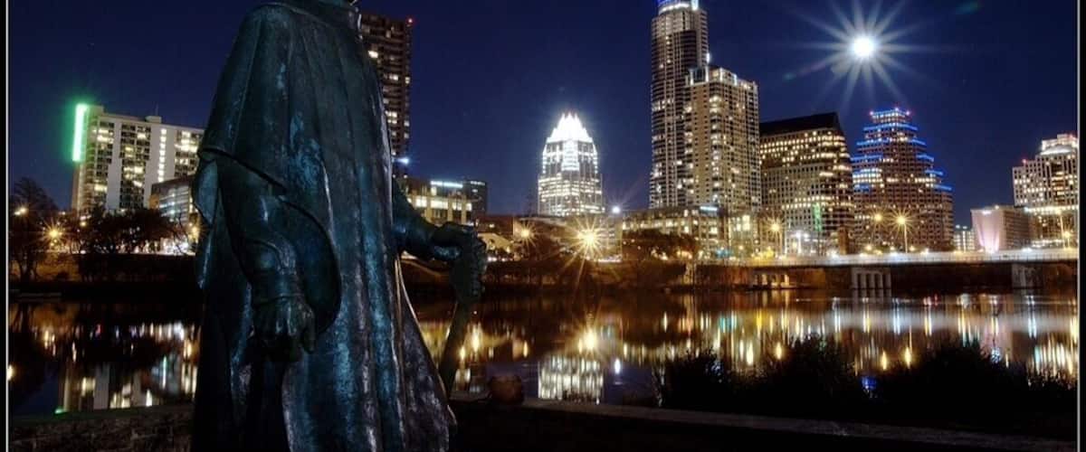A statue of a person in a hat by a body of water with a city skyline in the background.