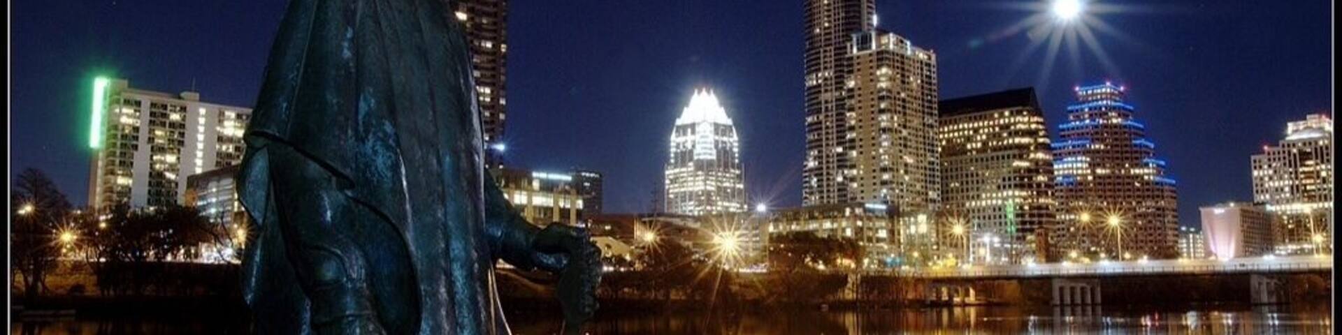 A statue of a person in a hat by a body of water with a city skyline in the background.