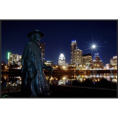 A statue of a person in a hat by a body of water with a city skyline in the background.