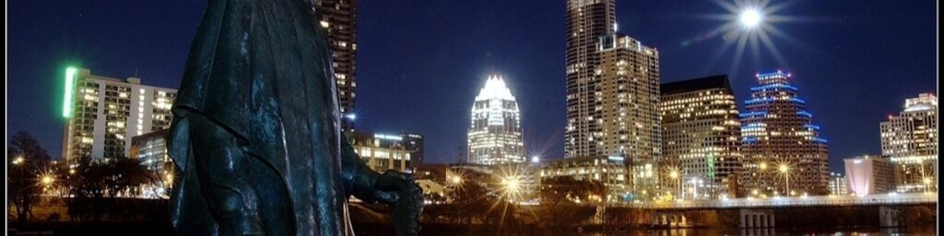 A statue of a person in a hat by a body of water with a city skyline in the background.