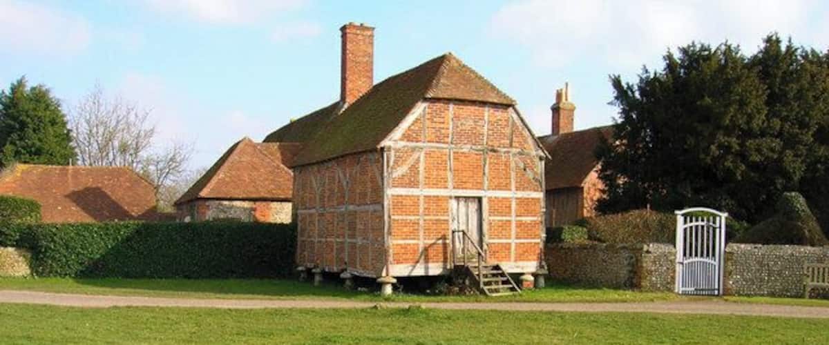 A small brick house with a sloped roof and a chimney, surrounded by a grassy field and a stone wall.