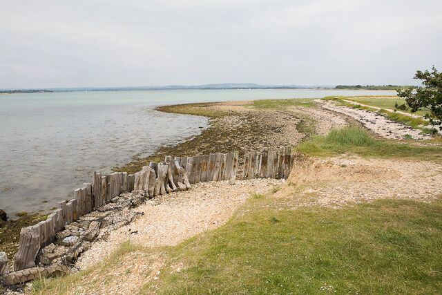 A coastal landscape with a wooden fence along a sandy beach, a grassy area, and a distant shoreline.