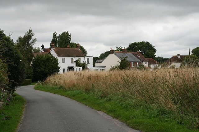 A narrow rural road curving through a village with white houses and tall grass.