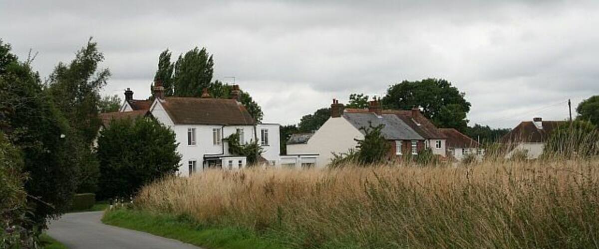 A narrow rural road curving through a village with white houses and tall grass.