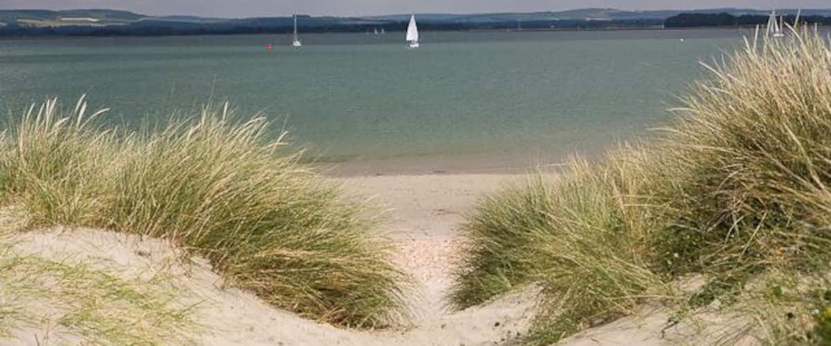 A beach with sand dunes and a calm lake with sailboats.