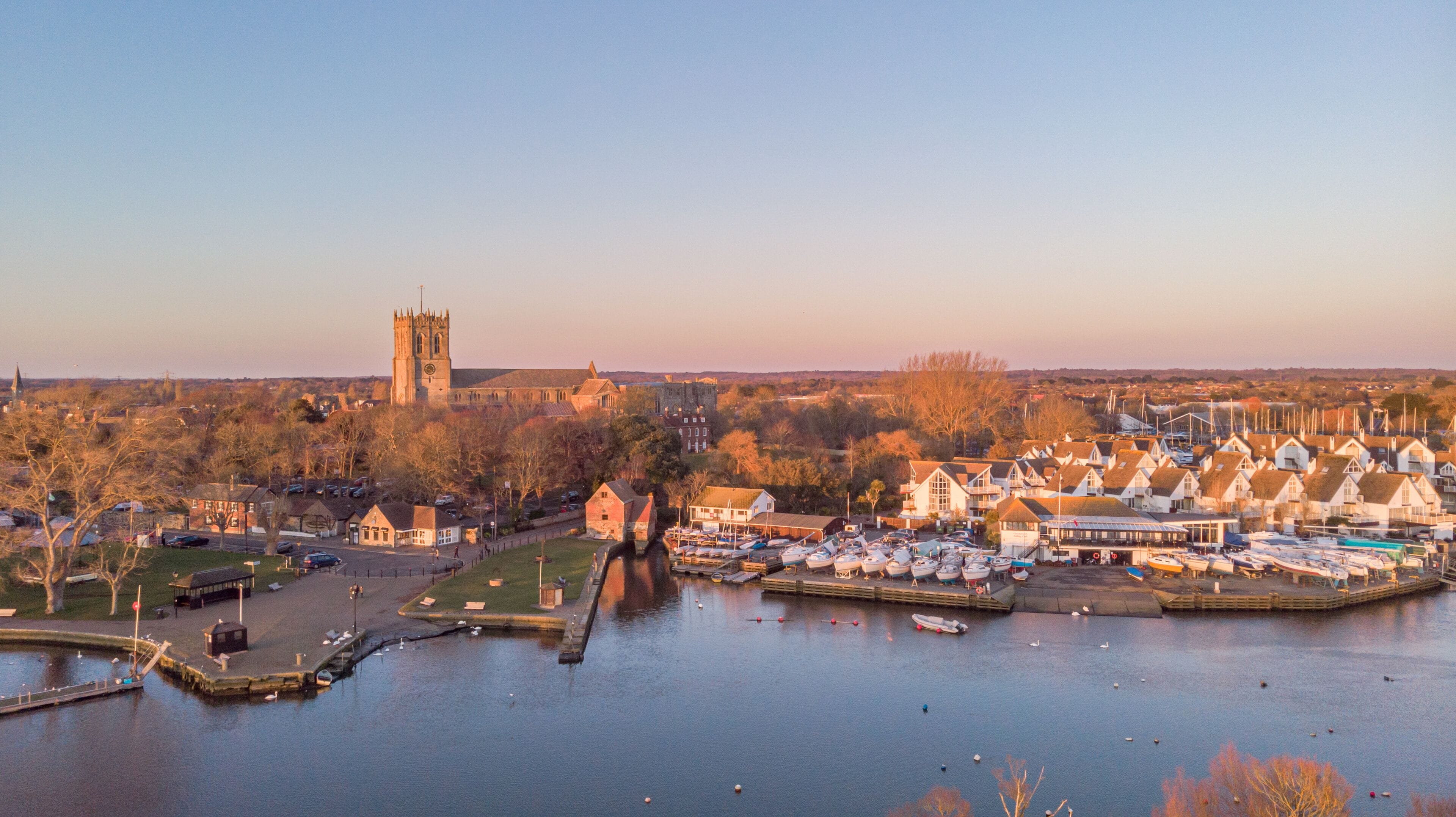 An aerial view of a river at golden hour with grassy bank and majestic church and marina in the background under a majestic blue sky