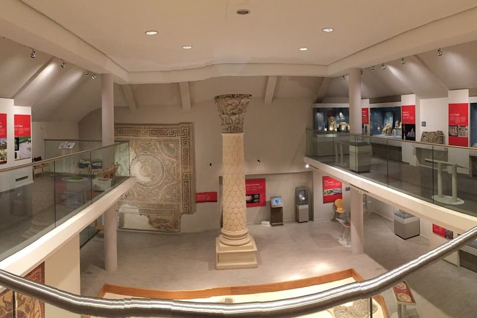 A museum interior with glass railings, a central column, and exhibits on the walls.