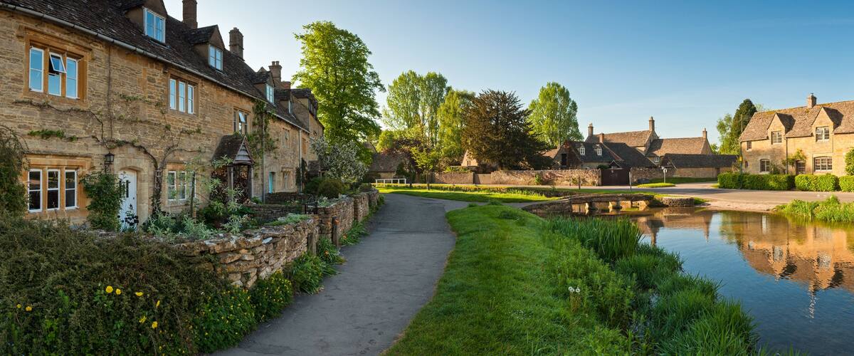 A serene riverside pathway flanked by historic stone buildings, a calm water body reflecting the architecture, and lush greenery.