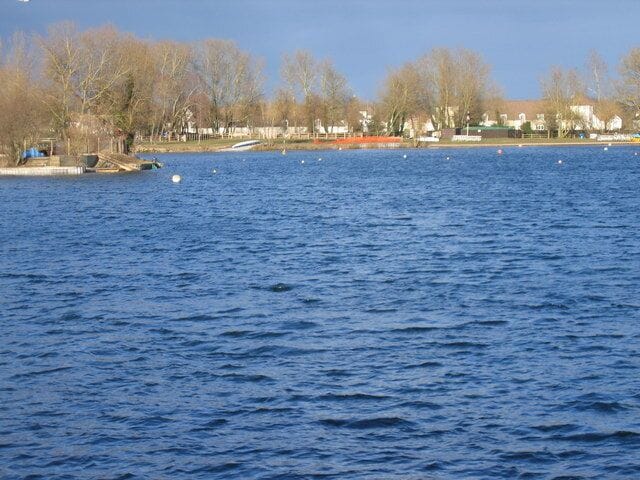 Un lago tranquilo con un muelle y barcos, rodeado de árboles sin hojas y casas al fondo.