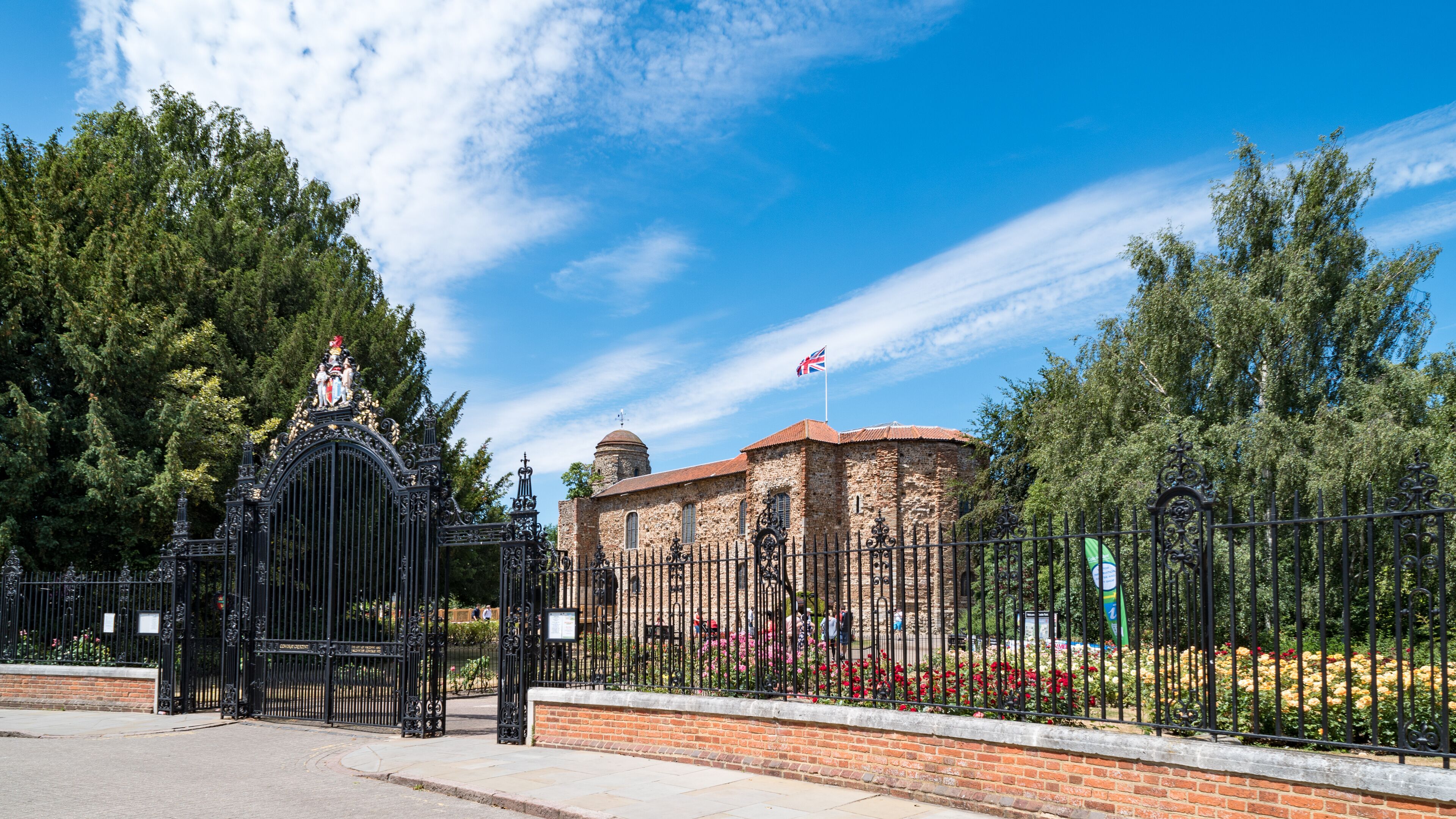 A historic building with a red-tiled roof, surrounded by a black wrought iron fence and a garden with red and yellow flowers.
