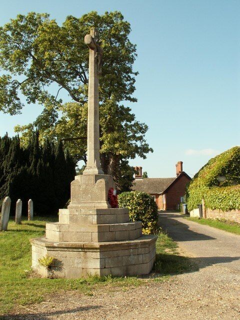 Um monumento de pedra com uma cruz no topo, rodeado por uma base circular, em ambiente rural com uma igreja e lápides ao fundo.
