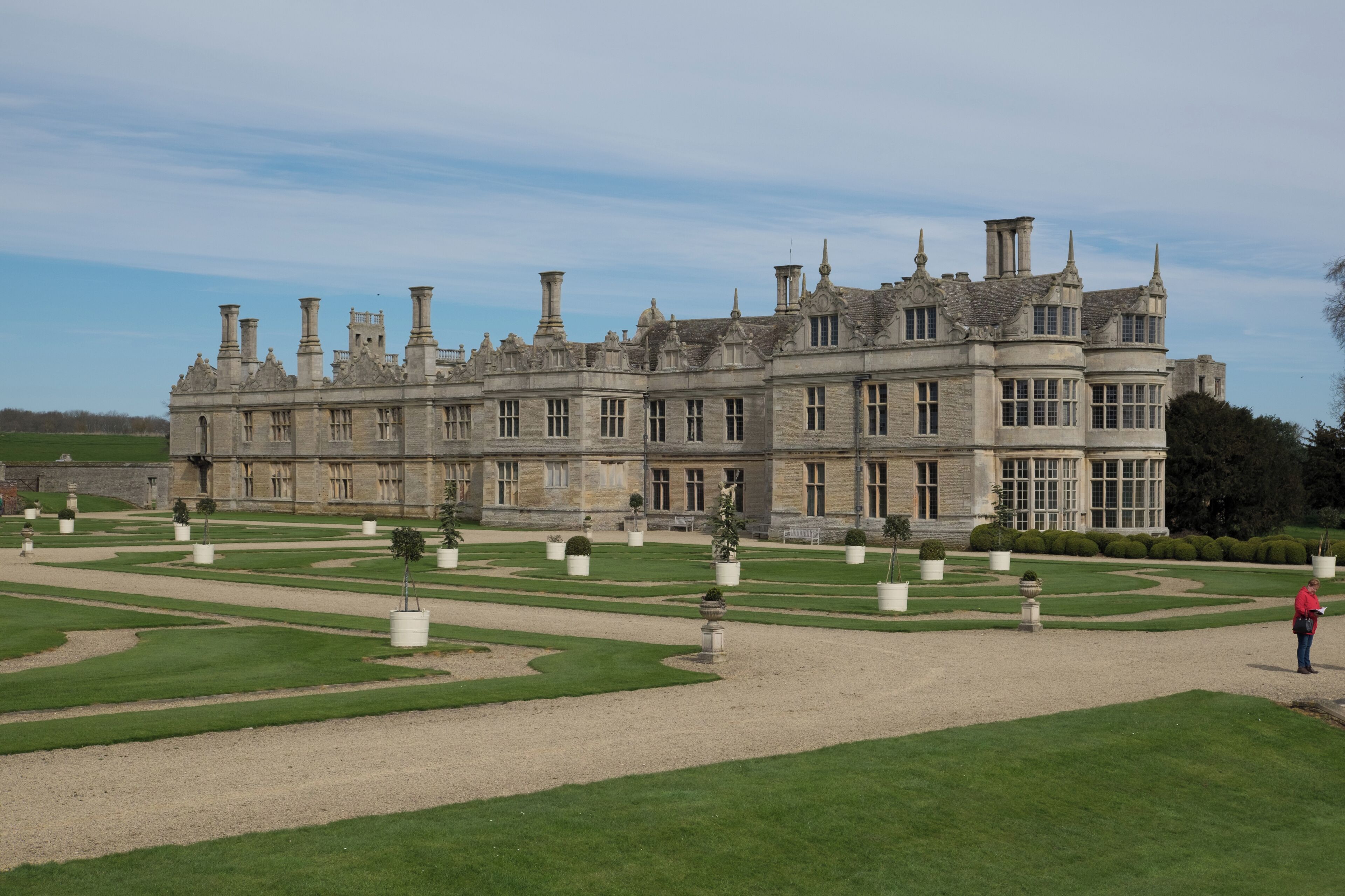 A large, historic building with a formal garden and a person standing in the foreground.