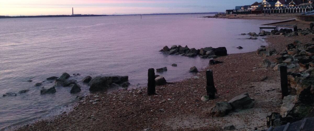 A pebbled beach with a pier and houses in the background.
