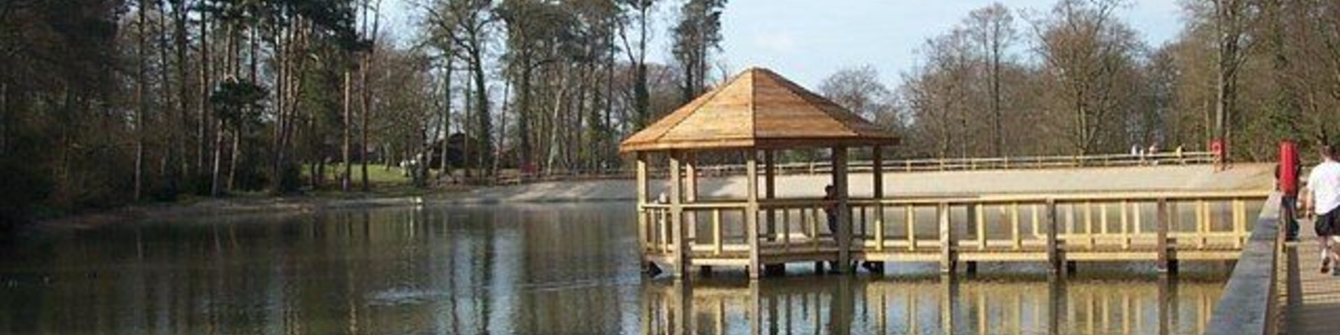Um gazebo de madeira à beira de um lago tranquilo, rodeado por árvores e um céu limpo.