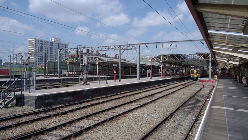 A train station platform with multiple tracks, overhead electrical lines, and a modern building in the background.
